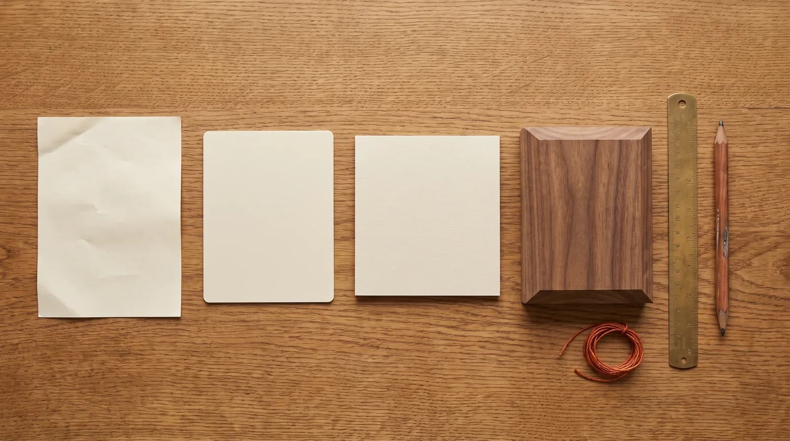 Four material samples on an oak table representing website price tiers: crumpled paper, plastic laminate, cardstock, and hand-finished walnut beside a brass ruler and pencil