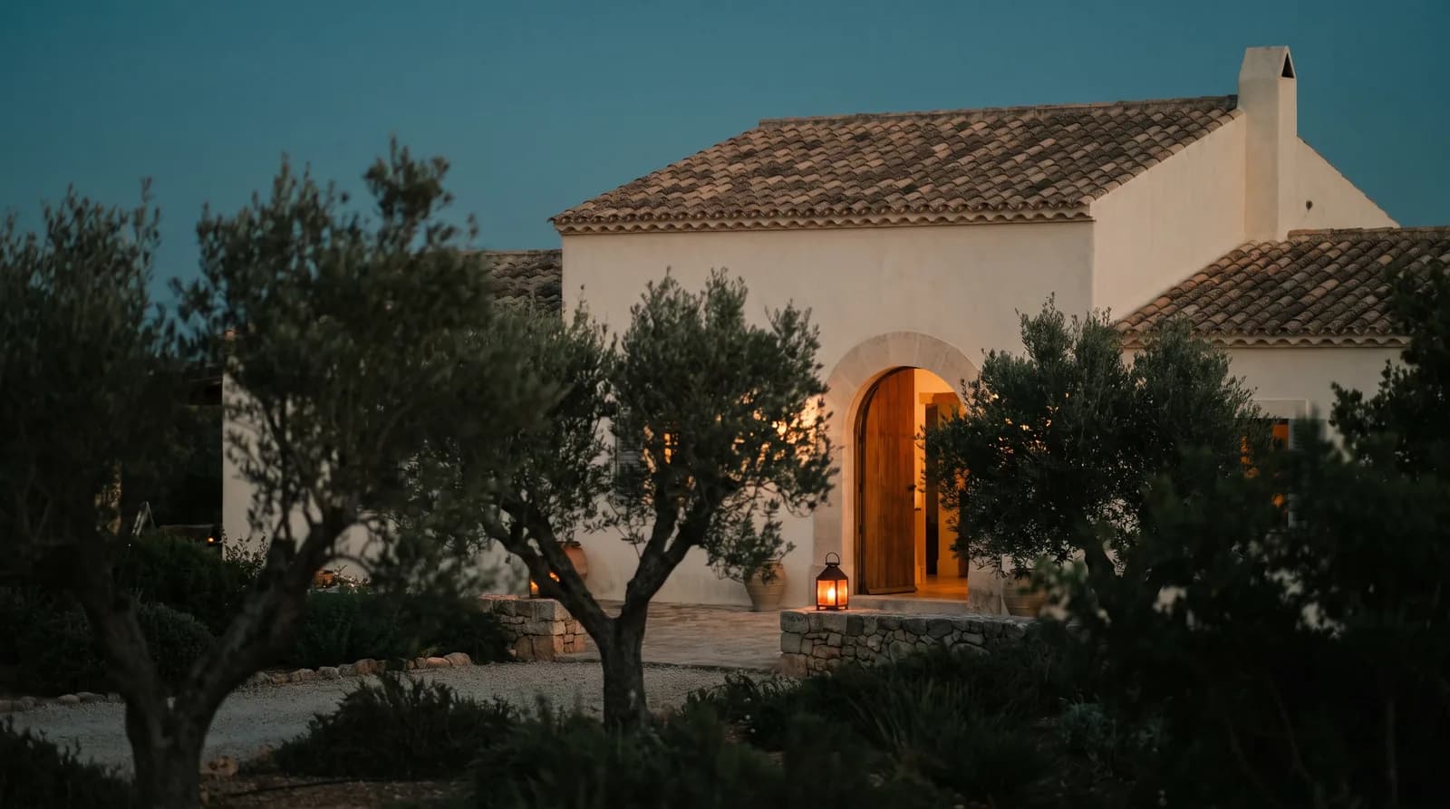 A whitewashed villa with a terracotta roof at twilight, its arched wooden door open and glowing from within, olive trees in the foreground