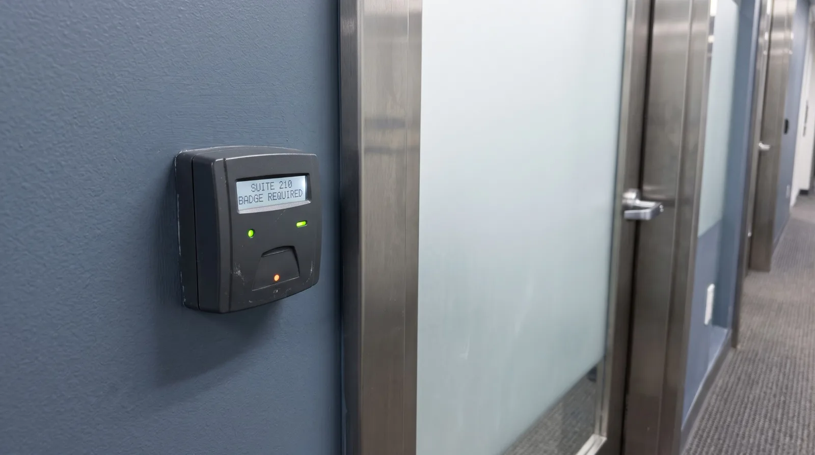 An access card reader on a slate blue corridor wall beside a frosted glass suite door, its screen reading SUITE 210, BADGE REQUIRED
