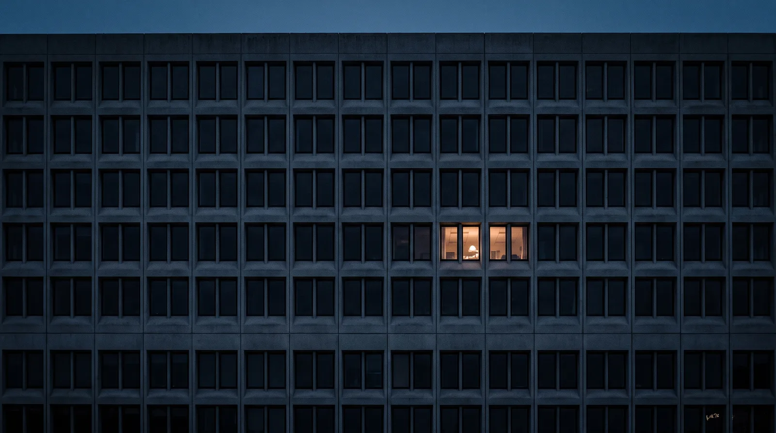 One lit office room, a desk lamp visible through its two windows, in the dense grid of an otherwise dark concrete facade at dusk