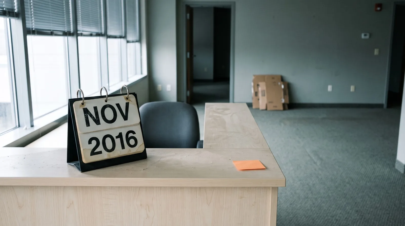 A black flip desk calendar with stained cards reading NOV 2016 on a dusty birch reception desk in an empty office suite, grey blinds on tall windows, flattened boxes against the far wall and a blank orange sticky note at the desk edge