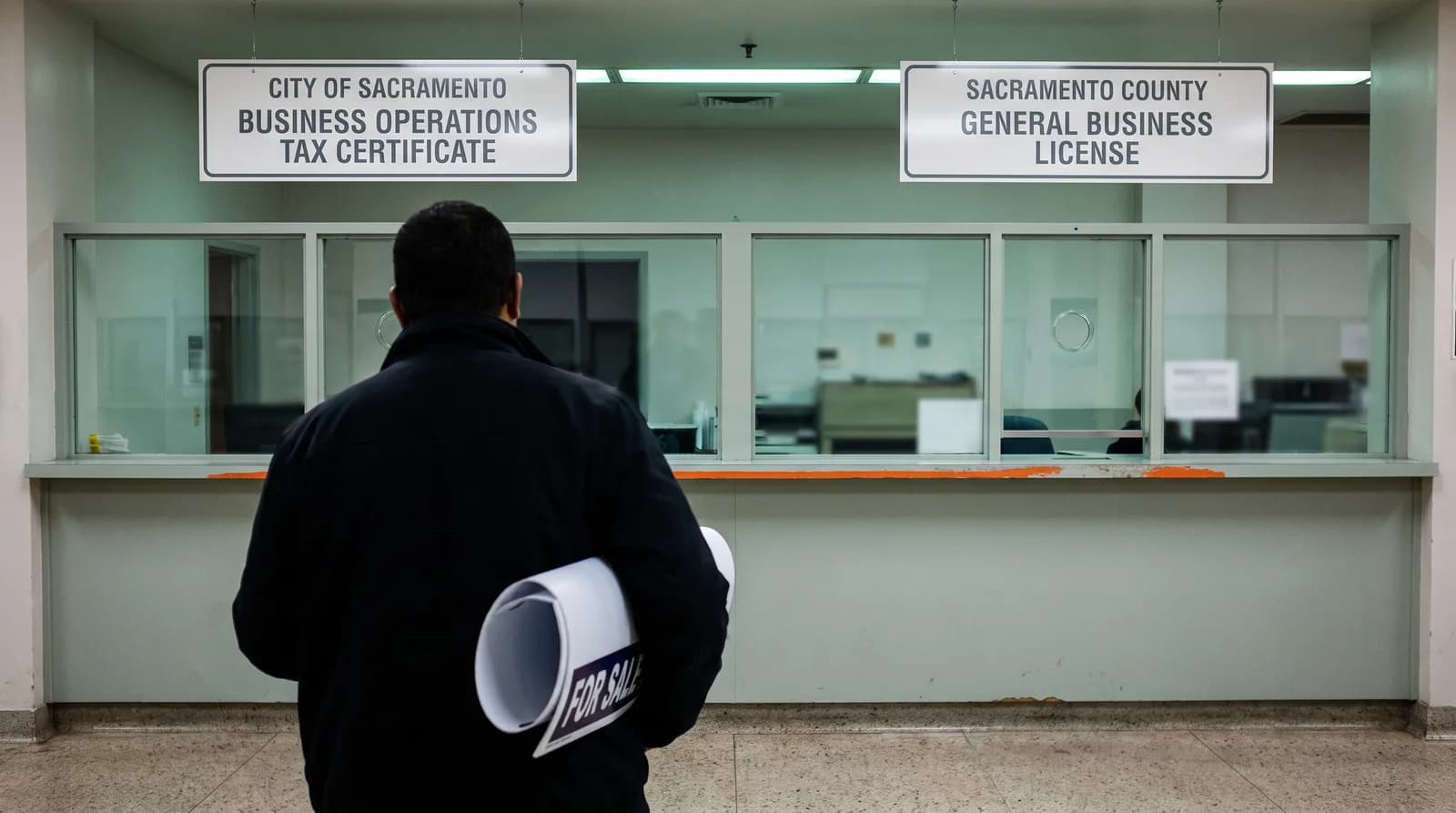 A person seen from behind at a government service counter, a rolled-up FOR SALE yard sign under one arm, standing between two overhead signs that read CITY OF SACRAMENTO, BUSINESS OPERATIONS TAX CERTIFICATE on the left and SACRAMENTO COUNTY, GENERAL BUSINESS LICENSE on the right