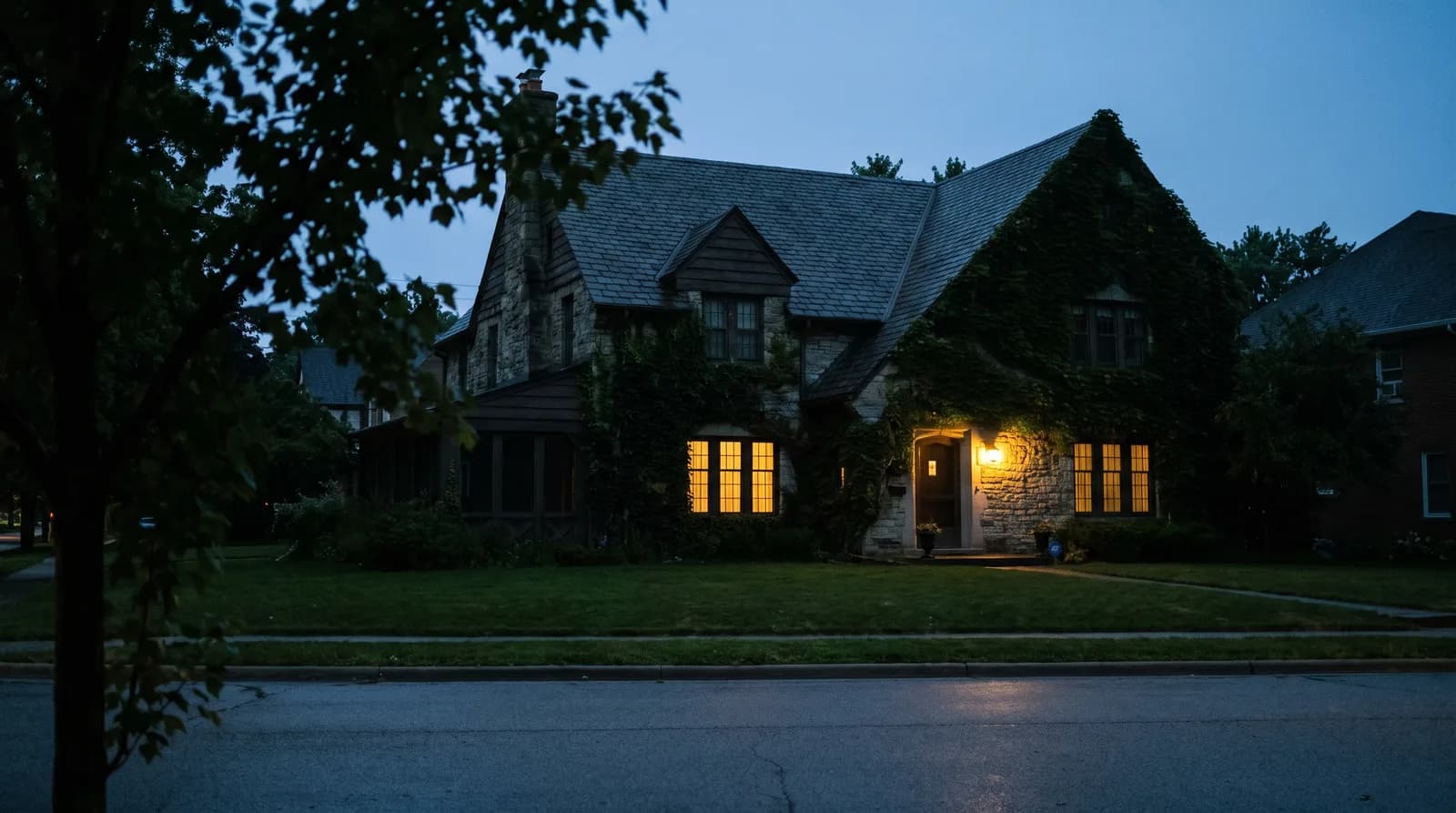 A house at dusk with warm windows and no for-sale sign on the lawn, seen from across a quiet street