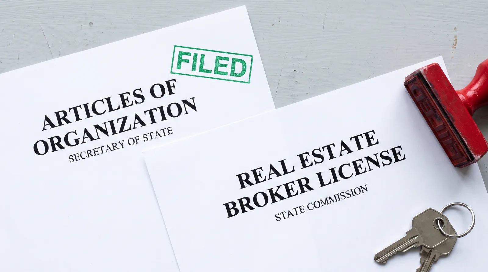 Two documents overlapping on a pale desk beside house keys, the left headed articles of organization, secretary of state and carrying a green FILED stamp, the right headed real estate broker license, state commission and left unstamped, with a red rubber stamp lying unused beside it
