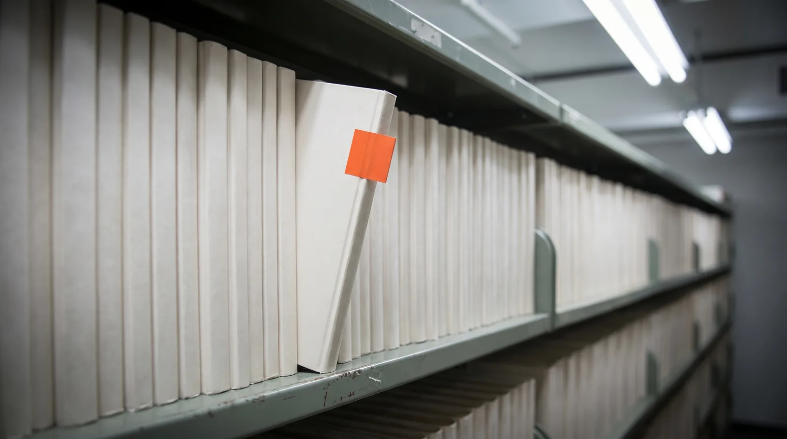 A long archive shelf of identical white bound volumes receding into blur under fluorescent light, with one volume pulled forward and tilted out, carrying a small orange tab