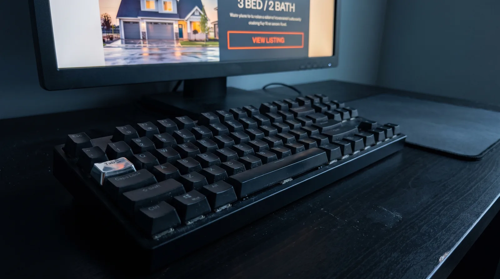 A keyboard on a dark desk whose Tab key is worn chrome-bright while every other key including Caps Lock is matte black, with no mouse and an empty mousepad, in front of a monitor showing a real estate listing headed 3 BED / 2 BATH whose VIEW LISTING button is wrapped in a bright orange keyboard focus outline
