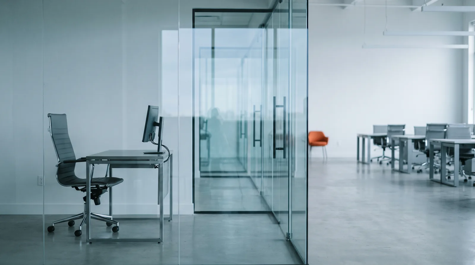 A single-desk private office behind a glass partition, with rows of empty desks on an open floor beyond and one orange chair against the far wall